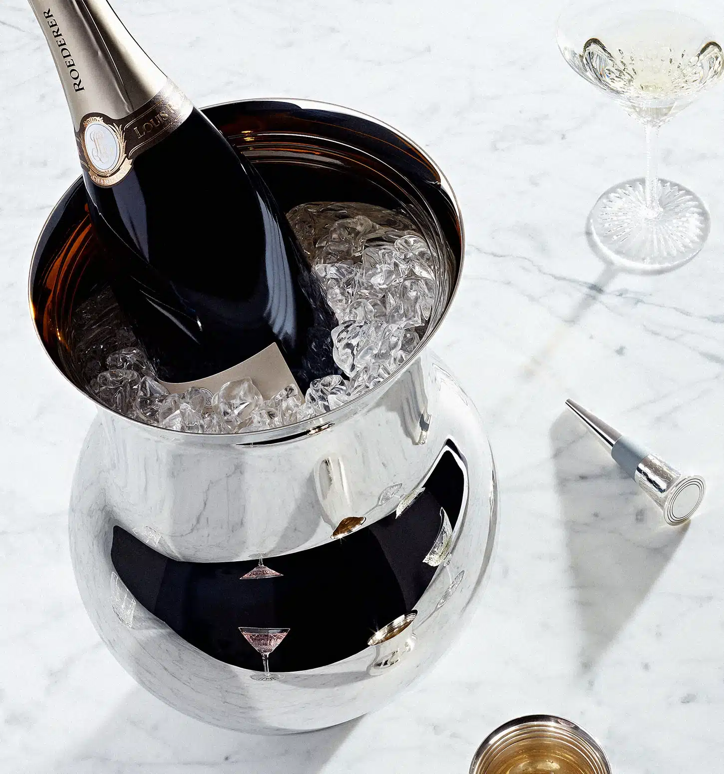 Champagne bucket in sterling silver from Pour le champagne collection full of ice cubes and a champagne bottle, photographed with a cristal glass, a champagne bottle stopper, on a marble table